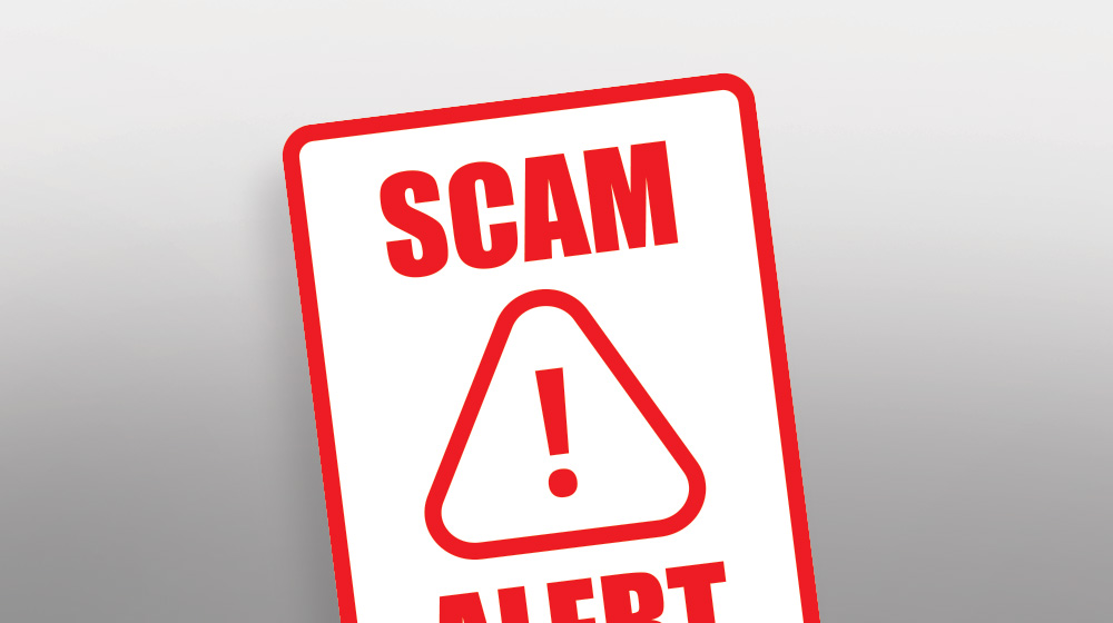 A white sign with red block letters that read SCAM ALERT. A triangle with an exclamation sign in the middle is positioned in the middle of the sign.