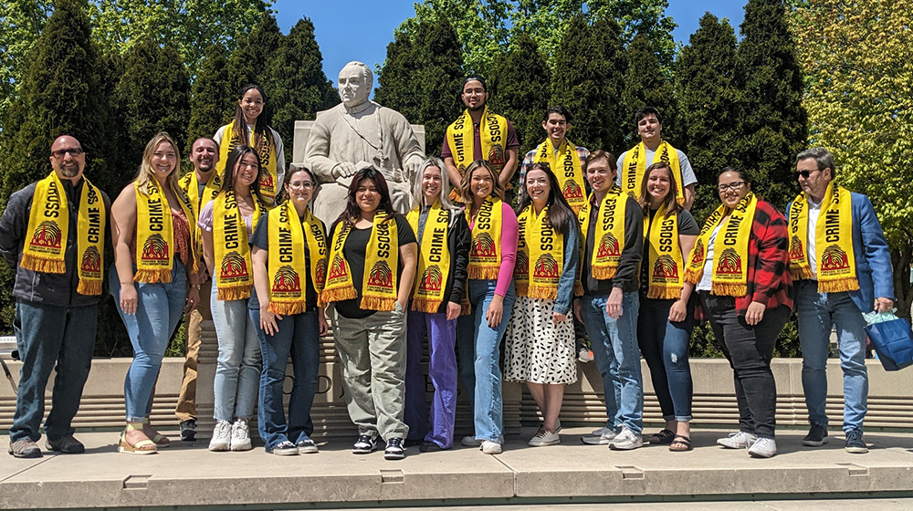 A large group of people gather for a photo in front of a statue. The people are all wearing yellow scarves with writing on them