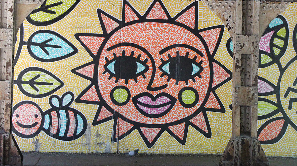 A mural of a mosaic sun on a wall under a train overpass