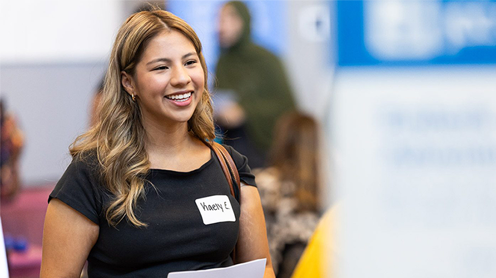 A young women student with brown hair, wearing a black blouse stands prepared to help the attendees at the Part-Time Job, Internship & Service Fair.